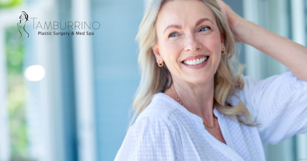 A smiling woman in a white shirt standing in front of a house.(MODEL) Tamburrino Plastic Surgery & Med Spa Logo