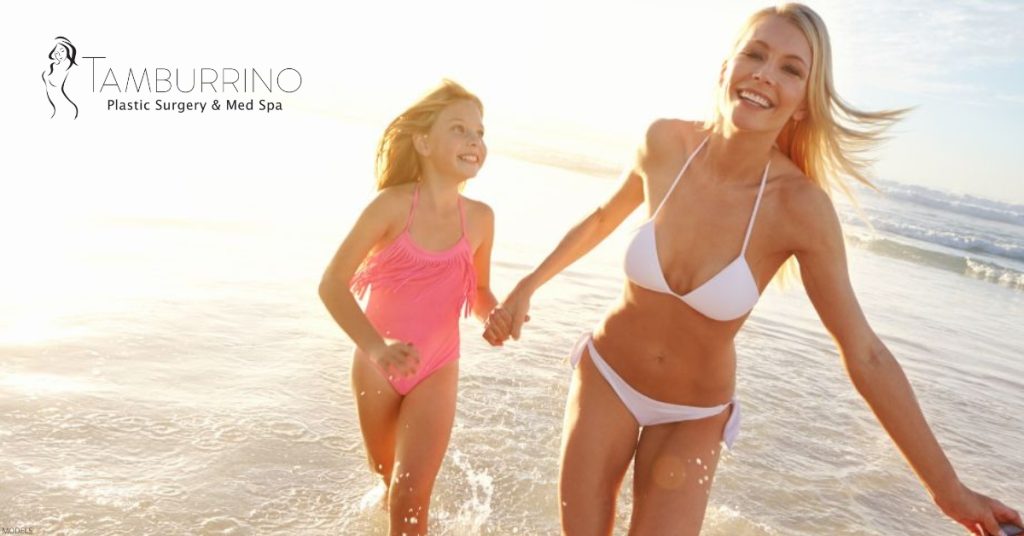 Mother and daughter running on the beach, smiling and enjoying the outdoors. (Model) Tamburrino Plastic Surgery & Med Spa logo displayed.